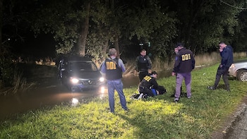 Noche, varios hombres en ropa de civil y chalecos DDI. Uno arrodillado junto a un detenido en el pasto, cerca de un vehículo negro semihundido en agua