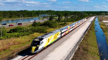 Tren de pasajeros Brightline de colores blanco, amarillo, naranja y rojo sobre vías, junto a una carretera con automóviles, un canal de agua, vegetación y árboles