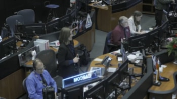 Vista aérea de una sala de control con varias personas sentadas en escritorios con múltiples monitores, y una persona de pie junto a una pantalla luminosa