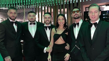 Cinco hombres con esmoquin y una mujer en vestido negro posan sonriendo en un salón con decoración de tubos luminosos verdes y piso de terrazo