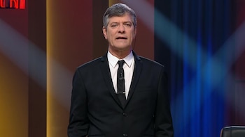 Nelson Castro, un hombre de mediana edad con cabello gris, viste un traje negro y corbata, hablando frente a un fondo oscuro con luces de colores ámbar y azul