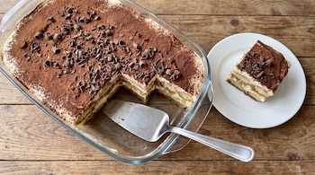 Un tiramisú en una fuente de vidrio con una espátula de metal, junto a una porción en un plato blanco sobre una mesa de madera.