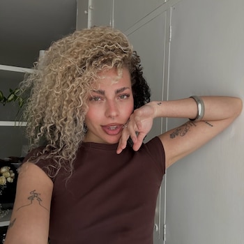Una mujer con cabello rizado de dos colores y tatuajes en los brazos posa frente a una pared blanca con una camiseta marrón