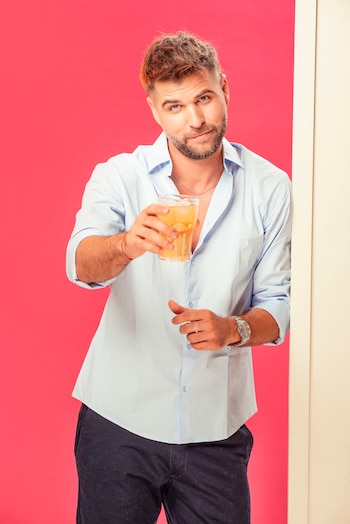 Hombre barbudo con camisa azul claro y pantalones oscuros sosteniendo un vaso con bebida y hielo, sonriendo levemente. Fondo rosa con borde crema a la derecha