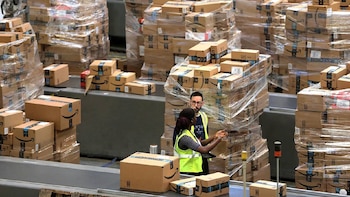 Dos trabajadores con chalecos reflectantes se encuentran entre pilas de paquetes de Amazon y cintas transportadoras en un centro de distribución. El hombre usa gafas
