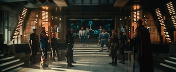Un grupo de superhéroes de pie dentro de una sala futurista con paneles luminosos, pantallas con gráficos y una plataforma elevada al fondo