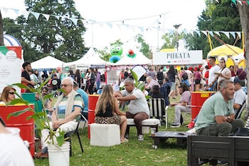 Festival al aire libre con multitud de personas en césped verde, puestos de comida, árboles, banderines y una mascota verde. Hay gente sentada y de pie