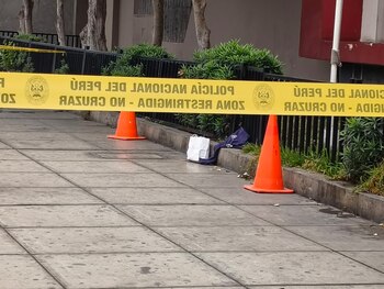 Una cinta amarilla de seguridad policial se extiende frente a dos conos naranjas, una bolsa blanca y una prenda azul en la acera