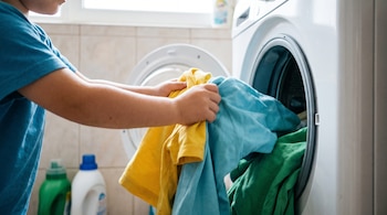 Manos de un niño meten ropa amarilla, celeste y verde en la lavadora blanca; se ven azulejos claros, una ventana y dos botellas de detergente.