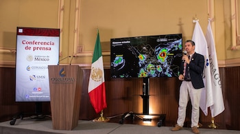 Un hombre habla con micrófono junto a una pantalla con un mapa de tormentas, un pódium de la Comisión Nacional del Agua y banderas.