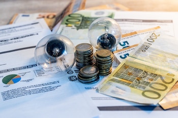 Sobre una mesa se ven varias facturas de la luz bajo varios billetes de euro, monedas y dos bombillas.