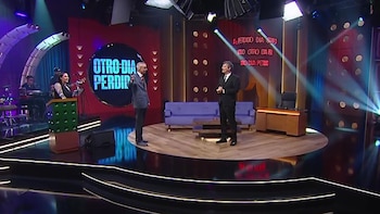 Vista amplia de un estudio de televisión moderno con dos hombres de traje conversando en el centro, una mujer detrás de un podio y músicos al fondo