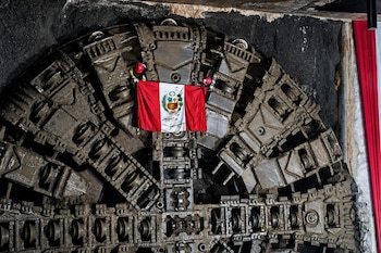 Vista frontal de la enorme cabeza de una tuneladora sucia y cubierta de lodo, con una bandera peruana izada en su parte central, dentro de un túnel oscuro
