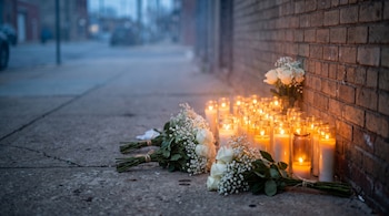 Varias veladoras encendidas y ramos de flores blancas colocados sobre una acera de concreto junto a una pared de ladrillos.