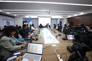 Sala de reuniones con personas sentadas en una mesa, computadoras portátiles, micrófonos, botellas de agua, banderas y cámaras de video