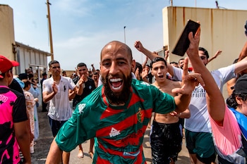 Un hombre barbudo con camiseta verde y roja sonríe con la boca abierta. Detrás, una multitud de hombres jóvenes mojados levanta los brazos. Edificios y cielo azul de fondo