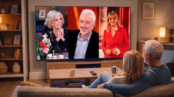 Un televisor con Mirtha Legrand, Andy Kusnetzoff y Teté Coustarot en pantalla. Una pareja de espaldas mira la televisión. Hay un sillón, mesa y control remoto.
