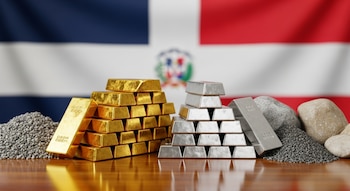 Lingotes de oro y plata apilados, pilas de grava y piedras sobre una mesa de madera pulida, ante una bandera de República Dominicana desenfocada.