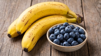 Dos bananas amarillas con manchas marrones y un tazón de cerámica lleno de arándanos sobre una superficie de madera.
