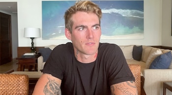 Un hombre rubio con camiseta negra y tatuajes visibles en el cuello y los brazos está sentado frente a un sofá y un cuadro de mar.