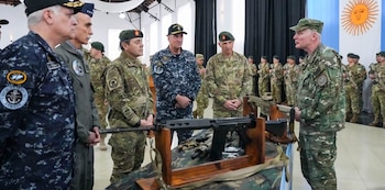 Militares en uniforme escuchan una presentación ante una mesa con fusiles sobre soportes de madera, junto a la bandera de Argentina