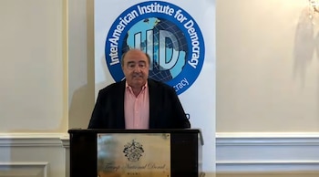 Hombre de cabello oscuro y saco oscuro de pie tras un podio con placa, frente a un banner blanco con el logo del InterAmerican Institute for Democracy