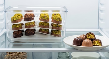 Bombones de chocolate con pistachos visibles, apilados en un contenedor transparente dentro de una heladera, con otros bombones en un plato blanco.