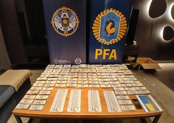 Billetes de cien dólares apilados ordenadamente sobre una mesa de madera, con dos pancartas azules de la Policía Federal Argentina (PFA) al fondo
