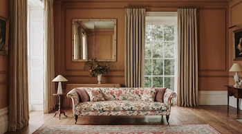 Salón con sofá estampado floral, dos mesas con lámparas, mueble consola, espejo, jarrón con flores, alfombra, dos ventanas y paredes paneladas.