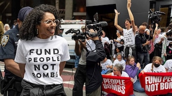 Una mujer sonriente con gafas y una camiseta "FUND PEOPLE NOT BOMBS" es escoltada por un oficial, mientras otros manifestantes sostienen pancartas