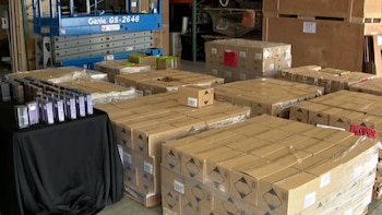 Imagen de almacén con numerosas pilas de cajas de cartón y madera, algunas con etiquetas de peligro. Una plataforma elevadora azul y una mesa con artículos pequeños