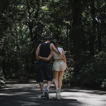 Un hombre y una mujer caminan de espaldas por un sendero asfaltado, rodeados de árboles y vegetación densa; el hombre abraza a la mujer
