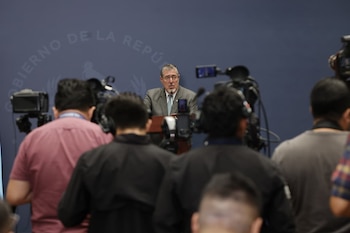 Un hombre con traje y gafas habla en un podio con micrófono frente a un fondo azul con el texto 'GOBIERNO DE LA REPÚBLICA'; periodistas y cámaras lo rodean