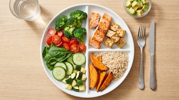 Un plato blanco dividido muestra salmón, tofu, brócoli, espinacas, tomates, pepino, calabacitas, arroz integral y camote. Al lado, un vaso de agua y aguacate.
