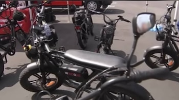 Uma visão de muitos veículos elétricos escuros de duas rodas, incluindo bicicletas e scooters com pneus grossos, exibidos no asfalto. Você vê uma tenda vermelha ao fundo