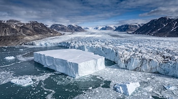 Bloque de hielo rectangular flotando en el agua frente a la pared de un glaciar con montañas rocosas al fondo.