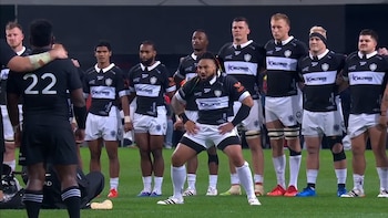Jugador Ma'a Nonu en uniforme blanco y negro ejecuta un haka. A su alrededor, otros jugadores de rugby con uniformes negros y bicolor de pie en el campo
