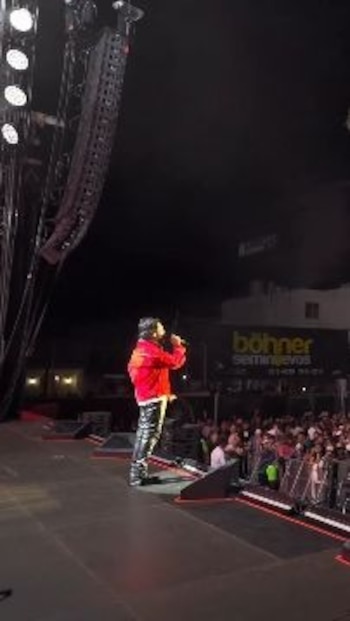 Un hombre de espaldas con chaqueta roja y pantalones negros está en un escenario con un micrófono. Se ven altavoces, público y un edificio con un letrero