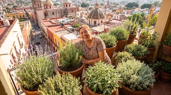 Mujer sonriendo rodeada de macetas con plantas de lavanda, romero, tomillo, salvia y santolina en un balcón. Al fondo, una ciudad con edificios y cúpulas.