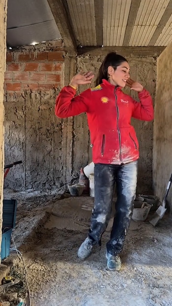 Mujer joven con chaqueta roja, pantalón oscuro y calzado manchados de cemento. Está en sala en construcción con paredes de ladrillo y hormigón, piso de tierra, cubos y herramientas