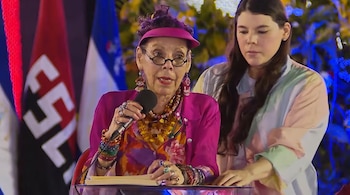 Rosario Murillo habla en micrófono, sostiene papeles en podio. Mujer joven detrás. Banderas, una roja con FSLN, y luces.