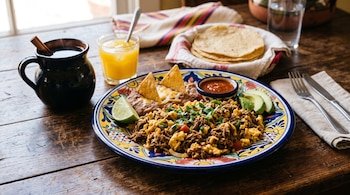 Plato con huevos revueltos y carne deshebrada, frijoles, totopos, salsa, aguacate y limón, junto a café, zumo y tortillas sobre madera.