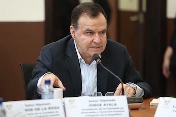 Primer plano de Jorge Ayala, un hombre con traje oscuro y camisa de rayas, hablando en un micrófono en una mesa de reuniones. Hay botellas de agua y placas de identificación en la mesa