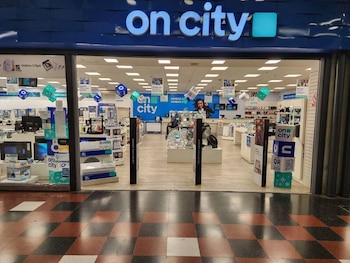 Fachada de la tienda On City con carteles promocionales, vidrieras, una puerta de entrada, ventiladores y televisores en exhibición. Piso con baldosas a cuadros