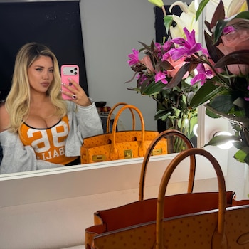 Mujer rubia tomando una foto a su reflejo en el espejo con un teléfono rosa, junto a un bolso naranja y flores