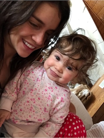 Selfie de una mujer sonriente con cabello oscuro y una niña pequeña de cabello rizado y ojos grandes, ambas mirando a la cámara