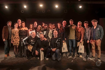 Grupo de veinticinco artistas y equipo de producción, sonrientes, posa en un escenario oscuro con focos. Algunos sostienen bolsas y certificados