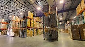 Amplio almacén con filas de estanterías metálicas llenas de cajas empaquetadas en palés, suelo de hormigón y techo con vigas expuestas e iluminación