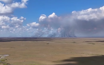 Vista aérea de una vasta llanura de pastizales marrones con una gran columna de humo gris elevándose en el horizonte bajo un cielo azul con nubes blancas