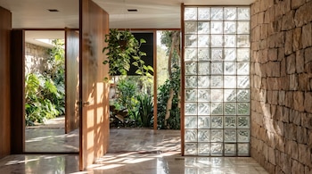 Vista de un pasillo con puertas de madera abiertas a un jardín, una planta colgante, una pared de bloques de vidrio y un muro de piedra rústica, con luz solar.